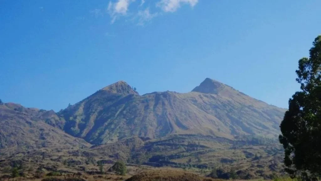 A scenic view of a twin-peaked mountain under a clear blue sky, with dry vegetation covering the slopes and a tree visible on the right side of the frame.