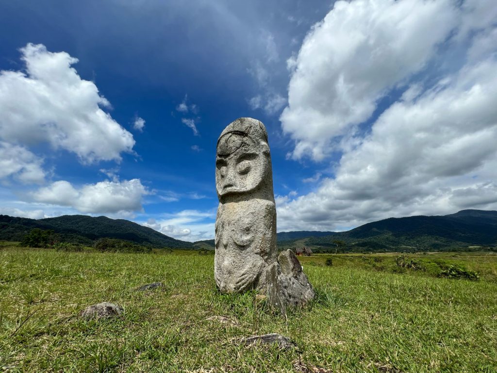 A lone stone statue standing in a grassy meadow with rolling hills and mountains in the background, under a bright blue sky with scattered clouds — a scene evoking adventure on a mountain trek.