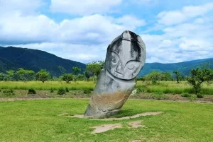 Alt text: An ancient megalith statue standing in a green valley with mountains in the background, a fascinating cultural sight discovered during a mountain trek in Central Sulawesi, Indonesia.