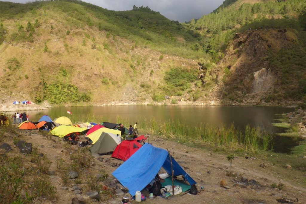 Colorful camping tents set up beside a serene lakeside surrounded by green hills, showcasing an adventurous outdoor experience for Sulawesi Jungle Tours.