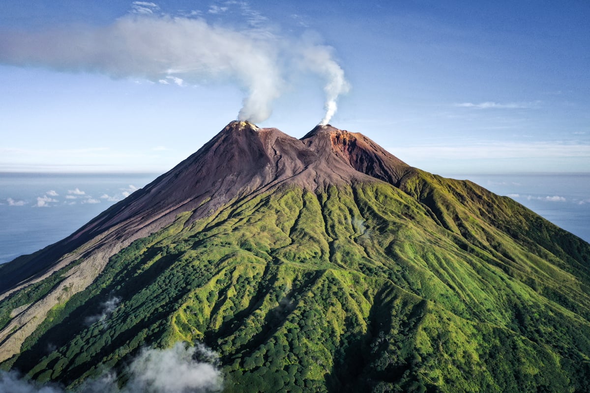 An active twin-peaked volcano surrounded by lush green valleys under a clear sky, showcasing the dramatic natural landscapes explored on Sulawesi Jungle Tours.