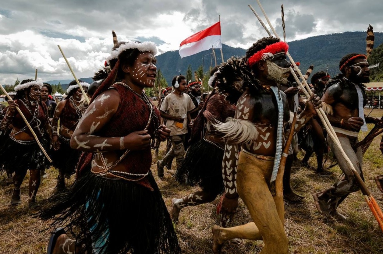 Papuan tribal warriors wearing traditional attire and body paint march together during a cultural performance at the Baliem Valley Festival 2026, with mountains and the Indonesian flag in the background.