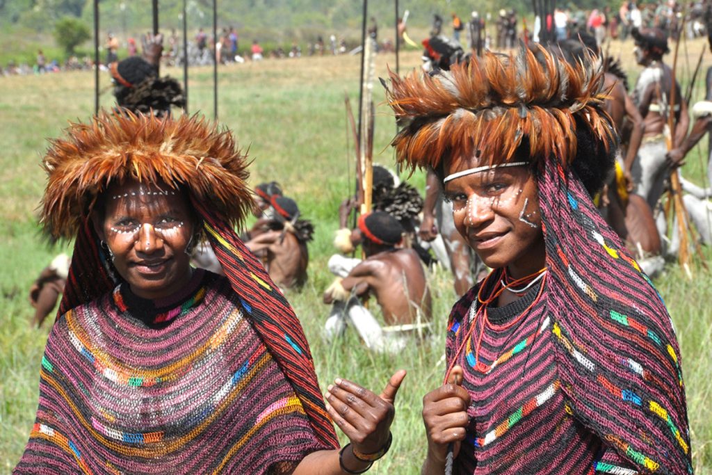 Two Papuan women wearing traditional feathered headdresses and woven garments during the Baliem Valley Festival 2026, with tribal warriors preparing for a cultural mock battle in the background.