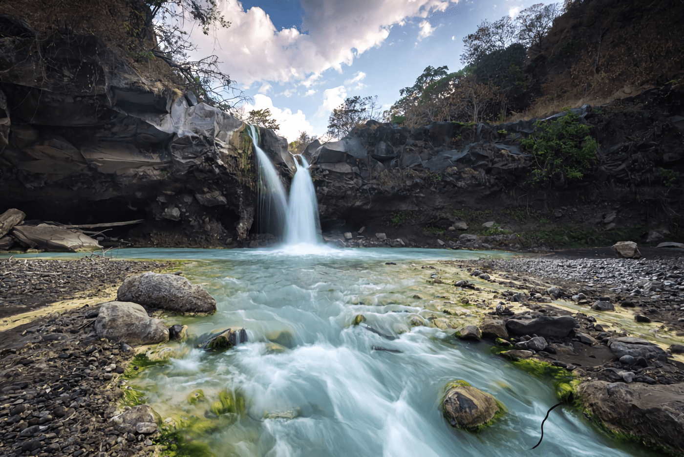 Hidden waterfalls in Lombok cascading between dark rocky cliffs into a clear turquoise pool, with flowing stream, scattered stones, and lush greenery under a bright blue sky.
